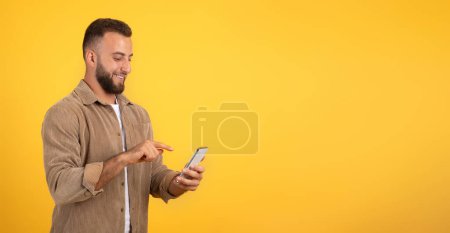 Photo for Positive european millennial guy with beard chatting on mobile phone, using app for blog, social networks, isolated on orange studio background, panorama. Work remotely, ad and offer - Royalty Free Image