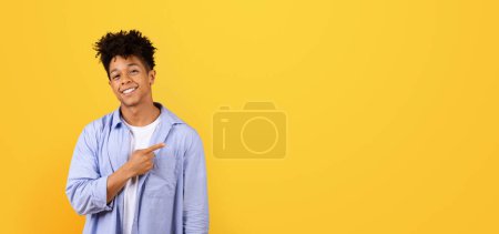 Téléchargez les photos : Homme noir en tenue élégante souriant et pointant vers le côté, mettant en valeur l'espace libre pour le texte ou le placement de produits sur fond de studio jaune vif, panorama - en image libre de droit