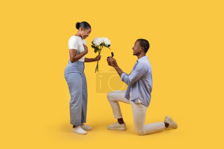 Photo for Black man on one knee holding an engagement ring towards surprised woman with bouquet of flowers against yellow background, depicting romantic proposal moment - Royalty Free Image
