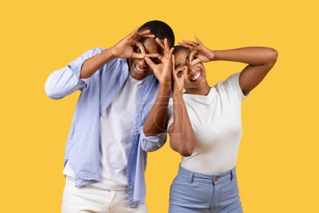 Téléchargez les photos : Homme et femme espiègles riant en faisant des lunettes avec leurs doigts, exprimant joie et créativité sur fond jaune vif dans un cadre décontracté - en image libre de droit