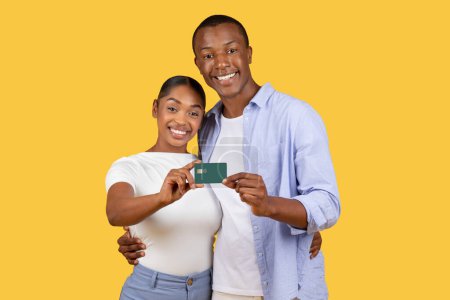 Téléchargez les photos : Souriant jeune couple noir présentant conjointement la carte bancaire, l'air heureux et content sur fond jaune solide, suggérant l'unité financière ou des dépenses partagées - en image libre de droit