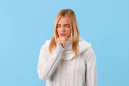 Photo for Woman in white sweater feeling unwell, coughing into her fist against plain blue background, with look of discomfort and sickness on her face - Royalty Free Image