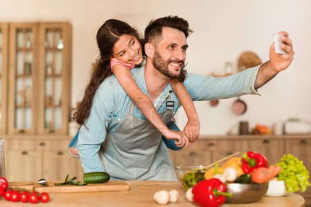 Photo for Delighted dad with his daughter on his back takes playful selfie in home kitchen brimming with colorful fresh produce, sharing special cooking moment together - Royalty Free Image