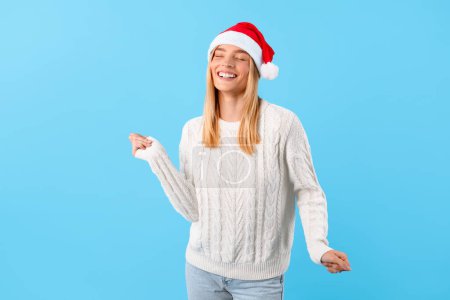 Photo for Blissful young lady wearing festive Santa hat and white sweater dancing and laughing against vivid blue backdrop, embodying the Christmas spirit - Royalty Free Image