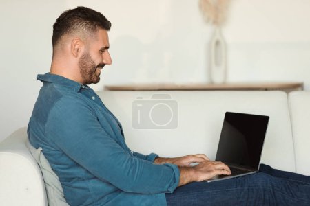 Photo for Side view of middle aged man smiles while working remotely on laptop as he sits on sofa in living room at home. Casual freelancer browses online, reflecting successful career - Royalty Free Image