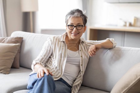 Photo for Portrait of beautiful senior woman relaxing on comfortable couch at home, happy elderly woman wearing eyeglasses sitting on sofa and looking at camera, enjoying retirement time, copy space - Royalty Free Image