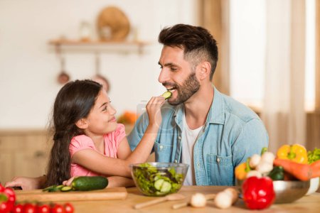 Téléchargez les photos : Joyeux papa partage tranche de concombre avec sa jeune fille pendant qu'ils préparent une salade verte fraîche dans l'intérieur de la cuisine accueillante, fille nourrir papa - en image libre de droit