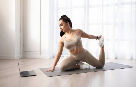 Photo for Online workout. Athletic well-fit beautiful brunette young woman in sportswear doing yoga or pilates on fitness mat at home, stretching legs, watching fitness video on pc laptop, copy space - Royalty Free Image