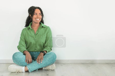Foto de La joven afro-americana positiva en camisa verde y jeans se sienta en el suelo, sonriendo mirando a un lado contra la pared blanca, encarnando un estilo de vida moderno y despreocupado. Espacio libre para anuncios de texto - Imagen libre de derechos