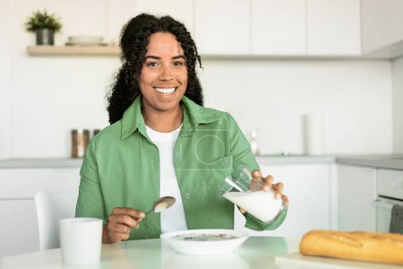Foto de Concepto de desayuno. Feliz mujer afroamericana vertiendo leche fresca de vidrio en un tazón con cereales, teniendo una deliciosa comida en el interior de la cocina moderna, sonriendo a la cámara - Imagen libre de derechos