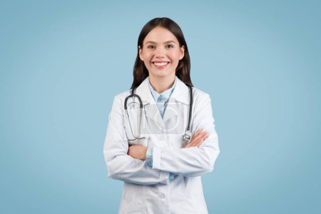 Confiada joven profesional médico en bata de laboratorio con estetoscopio brazos cruzados y sonriendo sobre fondo azul