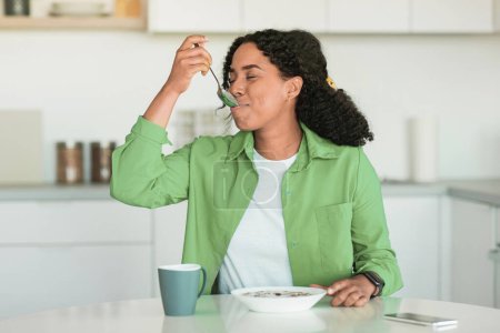 Photo for Black woman in green shirt starts her day with tasty breakfast in modern kitchen, sitting at dining table and savoring healthy cereals flakes and coffee, enjoys balanced meal and nutritious living - Royalty Free Image