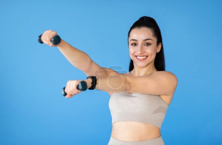 Photo for An active millennial woman with fit arms is exercising with small weights in a blue studio. Dressed in athletic wear, shes smiling at camera, representing her joy, toughness and determination - Royalty Free Image