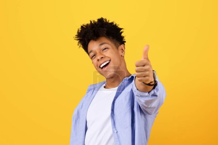 Photo for Excited young black male with curly hair flashes thumbs up sign, wearing casual shirt and smartwatch, exuding positivity against vibrant yellow backdrop - Royalty Free Image