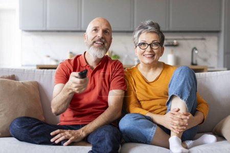 Photo for Happy Aged Spouses Watching Tv Together While Relaxing On Couch At Home, Smiling Senior Husband Aand Wife Resting On Comfortable Couch In Living Room, Man Switching Channels With Remote Controller - Royalty Free Image