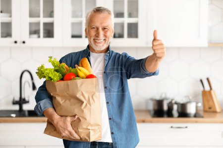 Photo for Happy Elderly Man Holding Paper Bag With Groceries And Showing Thumb Up, Cheerful Senior Gentleman Carrying Purchases And Smiling At Camera While Standing In Kitchen And Home, Free Space - Royalty Free Image