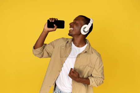 Téléchargez les photos : Joyeux jeune homme noir dans les écouteurs sans fil occasionnels chanter sur smartphone mobile avec écran vide, profiter de temps libre, amusant, isolé sur fond de studio jaune. Annonce et offre, application pour karaoké - en image libre de droit