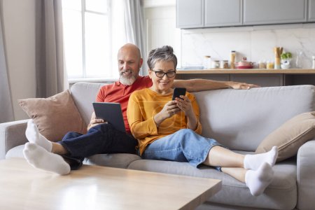 Téléchargez les photos : Vieux couple souriant reposant sur un canapé avec tablette numérique et téléphone portable, heureux homme et femme âgés utilisant les technologies modernes à la maison, se détendre avec des gadgets, profiter des loisirs de retraite - en image libre de droit