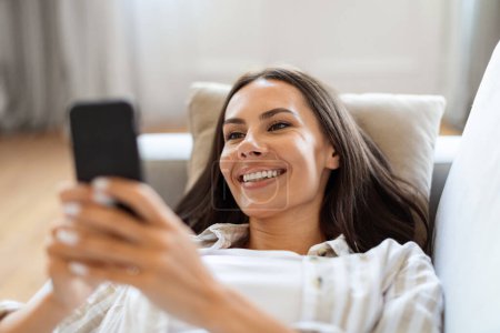 Photo for Young Smiling Woman Using Smartphone While Resting On Sofa At Home, Beautiful Millennial Female Lying On Couch With Mobile Phone, Messaging With Friends Or Browsing New App, Closeup - Royalty Free Image