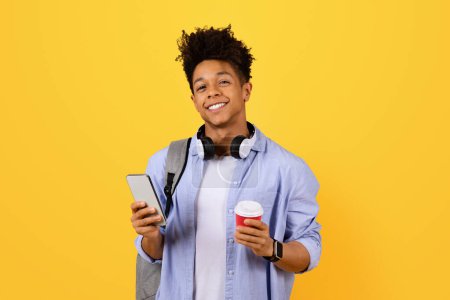 Foto de Hombre negro sonriente sosteniendo teléfono inteligente y taza de café, con auriculares alrededor del cuello, correa de la mochila visible, en postura relajada contra el fondo amarillo - Imagen libre de derechos