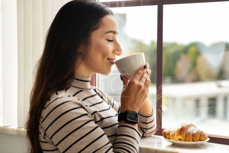 Téléchargez les photos : Souriant jeune femme arabe boire une tasse de café chaud, thé avec croissant dans le salon léger, cuisine intérieure près de la fenêtre. Dame profiter de temps libre, calme, paix et petit déjeuner et style de vie confortable à la maison - en image libre de droit