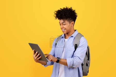 Photo for Stylish black man as student using digital tablet while wearing backpack, standing against yellow studio background, embodying modern education - Royalty Free Image