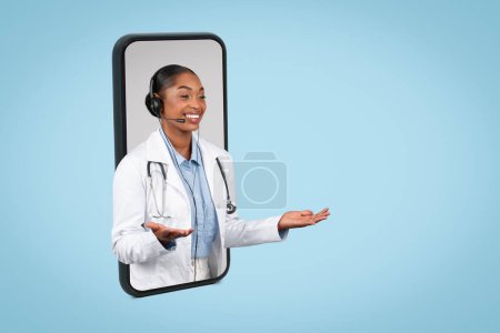 Téléchargez les photos : Heureux médecin femme noire millénaire dans les écouteurs tenir de l'espace libre sur les mains, écarter les bras sur grand écran de smartphone, isolé sur fond bleu, studio. Aide médicale à distance - en image libre de droit