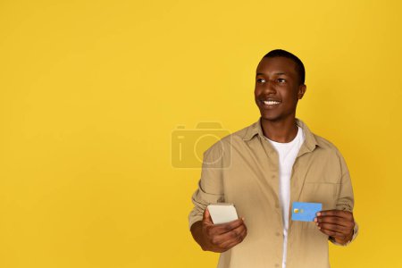 Photo for Cheerful pensive young black man use mobile phone and credit card, dream, think, look at free space, isolated yellow studio background. App for banking and sale, online shopping, pay money - Royalty Free Image