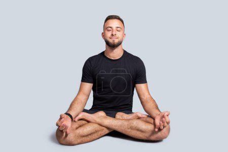 Foto de Hombre europeo milenario positivo con barba en ropa deportiva practica yoga en posición de loto, disfruta de ejercicios de respiración, calma, paz y cuidado de la salud, aislado sobre fondo gris estudio - Imagen libre de derechos