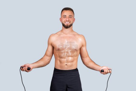Photo for Positive sweat strong muscular millennial european guy with naked torso make exercises with skipping rope, enjoy cardio workout, isolated on gray studio background. Sport athlete, weight loss - Royalty Free Image