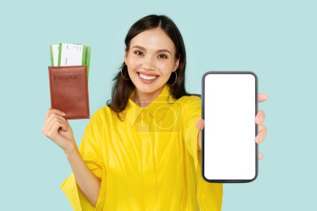 Photo for Positive pretty brunette young woman tourist holding passport with travel tickets and smart phone with mockup empty screen, standing on isolated blue background, great travelling offer, online checkin - Royalty Free Image