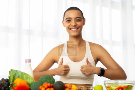 Photo for Smiling millennial latin woman in sportswear make thumb up sign with hands in kitchen interior, recommends fresh vegetables, salad. Diet fit, weight loss, healthy food and lifestyle approve at home - Royalty Free Image