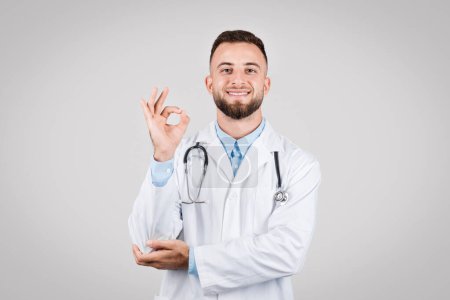 Photo for Confident caucasian male doctor in lab coat with stethoscope around neck gesturing okay sign with hand, expressing approval or success, grey background - Royalty Free Image