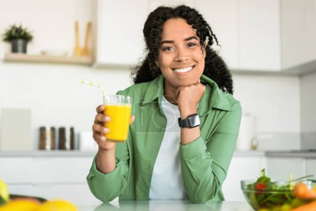 Téléchargez les photos : Heureuse femme noire tient un verre de jus d'orange et sourit à la caméra dans sa cuisine intérieur moderne. Portrait de dame aime boisson de désintoxication et petit déjeuner symbolisant un nouveau départ à sa journée - en image libre de droit