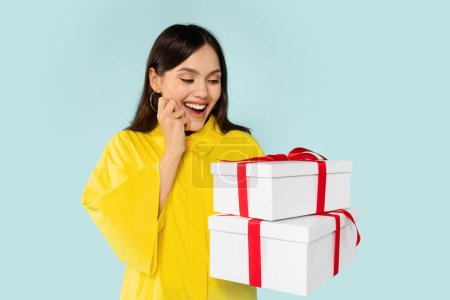 Photo for Excited pretty young woman looking at presents white gift boxes with red bow in her hand and grimacing, happy lady got birthday or anniversary gifts, isolated on blue background, copy space - Royalty Free Image