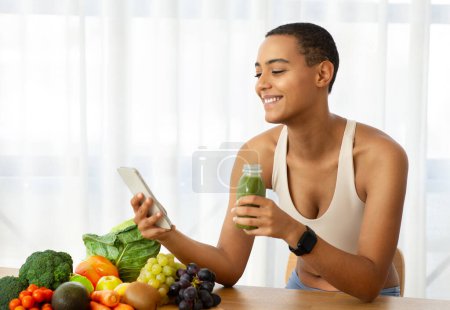 Photo for Happy millennial latin woman in sportswear, drink jar of fresh smoothie, juice, watch on smartphone on kitchen interior with fresh vegetables. Diet, weight loss at home, healthy food, app blog - Royalty Free Image