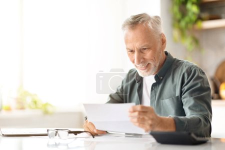 Téléchargez les photos : Gentleman âgé souriant lisant des documents alors qu'il était assis au bureau dans la cuisine, homme âgé heureux vérifiant le courrier à la maison, examinant des documents de la compagnie d'assurance, gros plan avec de l'espace de copie - en image libre de droit