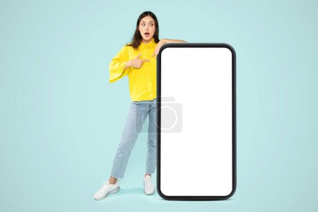 Photo for Amazed pretty stylish millennial woman pointing at huge phone with white blank screen over blue studio background, mockup, copy space for advertisement, mobile app, online offer - Royalty Free Image