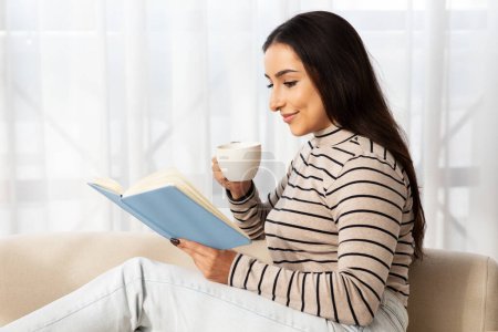 Foto de Mujer árabe joven calma bonita leer libro, trabajo de planificación, sentarse en el sofá, beber taza de café en el interior de la sala de estar luz. Señora disfrutar del tiempo libre, estudio, educación, afición y estilo de vida acogedor en casa - Imagen libre de derechos