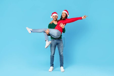 Foto de Alegre pareja europea en los sombreros de Santa divertirse mientras posan en el estudio, el hombre lleva a la mujer celebrando las vacaciones de Navidad y Año Nuevo sobre el telón de fondo azul. Longitud completa, espacio de copia - Imagen libre de derechos