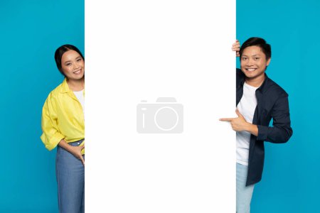 Photo for Cheerful young chinese family in casual recommend information on big banner with empty space for words, text, ad and offer, isolated on blue studio background. Good news, attention sale - Royalty Free Image