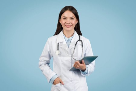 Téléchargez les photos : Jeune médecin confiante avec stéthoscope autour du cou tenant une tablette numérique, prête pour les soins aux patients, sur fond bleu - en image libre de droit