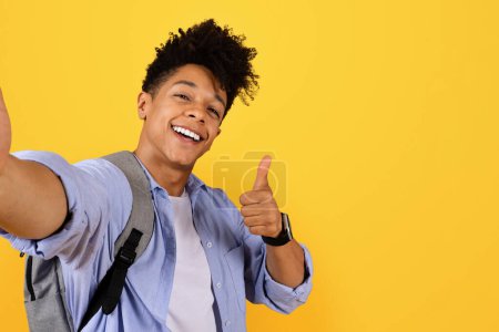 Foto de Feliz estudiante negro con mochila tomando selfie y dando pulgares hacia arriba signo, con expresión alegre, sobre el fondo amarillo espacio libre - Imagen libre de derechos