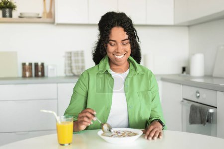 Téléchargez les photos : Joyeuse dame noire aime les céréales avec du lait pour le petit déjeuner assis dans la cuisine domestique. Femme décontractée en chemise verte mangeant un repas sain le matin. Nutrition et recettes pour votre santé - en image libre de droit