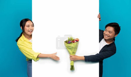 Téléchargez les photos : Homme asiatique millénaire souriant dans occasionnel donner bouquet de fleurs à la femme sur une grande bannière avec espace vide, isolé sur fond de studio bleu. Date, anniversaire romantique, anniversaire félicitations - en image libre de droit