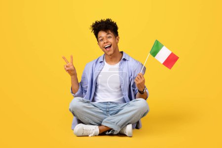 Photo for Joyful young black male student in casual clothes sitting cross-legged on yellow background, flashing peace sign while holding the flag of Italy with bright smile - Royalty Free Image