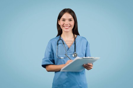 Confident female healthcare professional in scrubs holding clipboard and wearing stethoscope, standing with friendly smile on blue background