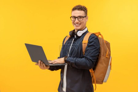 Foto de Feliz joven guapo en traje casual llevando mochila estudiante usando el ordenador portátil en el fondo del estudio amarillo, disfrutando de clases en línea. Concepto de educación electrónica - Imagen libre de derechos