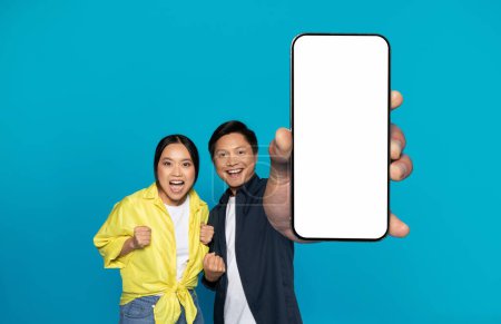 Photo for Glad excited young chinese couple in casual celebrate win, success, show smartphone with empty screen, isolated on blue studio background. Recommendation gadget, ad and offer, online sale, shopping - Royalty Free Image