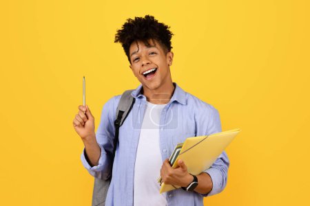 Téléchargez les photos : Joyeux étudiant noir tenant dossiers et stylo, riant et regardant excité, portant un sac à dos et smartwatch, ensemble sur fond jaune vibrant - en image libre de droit